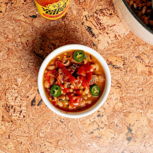 A white bowl full of Jalapeño black-eyed peas, topped with Yellowbird Jalapeño Hot Sauce and sliced green jalapeños. The hot sauce bottle is in the background.