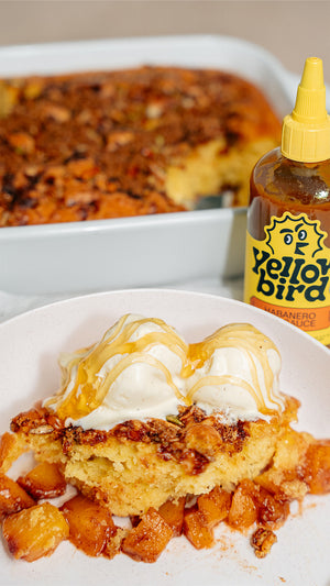 A piece of cake topped with ice cream and honey in the foreground, with a bottle of Yellowbird Habanero Hot Sauce and the rest of the cake in the background.