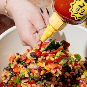 Cowboy Caviar loaded onto a tortilla chip over the bowl of Cowboy Caviar. Extra Yellowbird Sriracha is being drizzled on the loaded chip from a squeeze bottle.