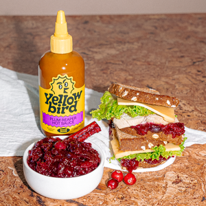 A dish of Plum Reaper cranberry sauce next to a turkey sandwich and a bottle of Yellowbird Plum Reaper Hot Sauce.