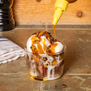  A clear glass dish of vanilla ice cream with sautéed plums, bacon bits, and a drizzle of Yellowbird Plum Reaper Hot Sauce.