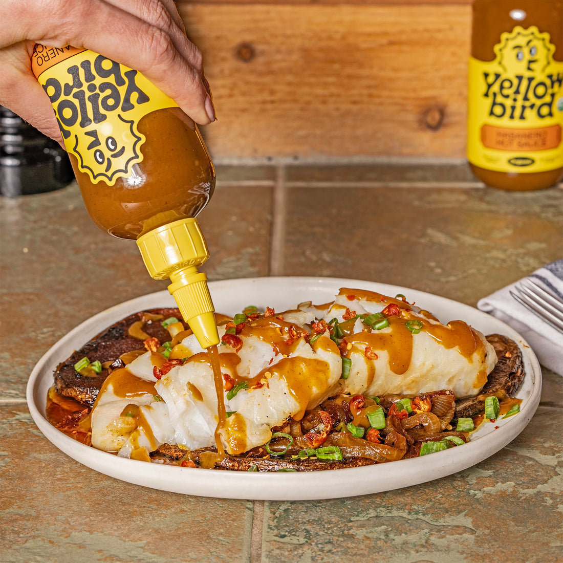 A plate of cooked cod over caramelized onions, with Yellowbird Organic Habanero Hot Sauce being drizzled on top.