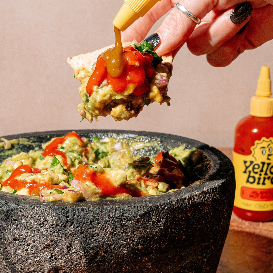 Guacamole in a stone mortar, drizzled with Yellowbird Jalapeño Hot Sauce. A hand holds a chip above, loaded with guac, and drizzling Yellowbird Serrano Hot Sauce onto the chip. There is a bottle of Yellowbird Jalapeño Hot Sauce in the background. 