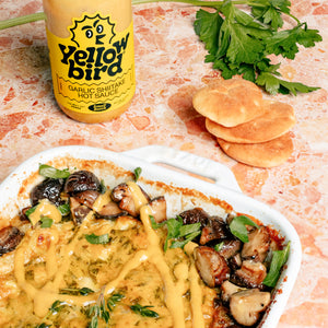 Baked brie with mushrooms and herbs, topped with a drizzle of Garlic Shiitake Hot Sauce, next to a bottle of the hot sauce and mini pita breads. 