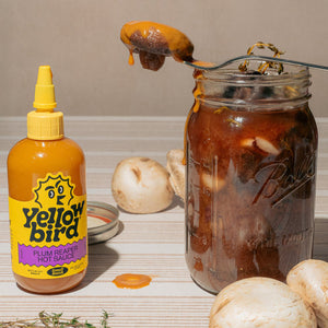 A bottle of Yellowbird Plum Reaper Hot Sauce next to a mason jar filled with spicy pickled mushrooms. 