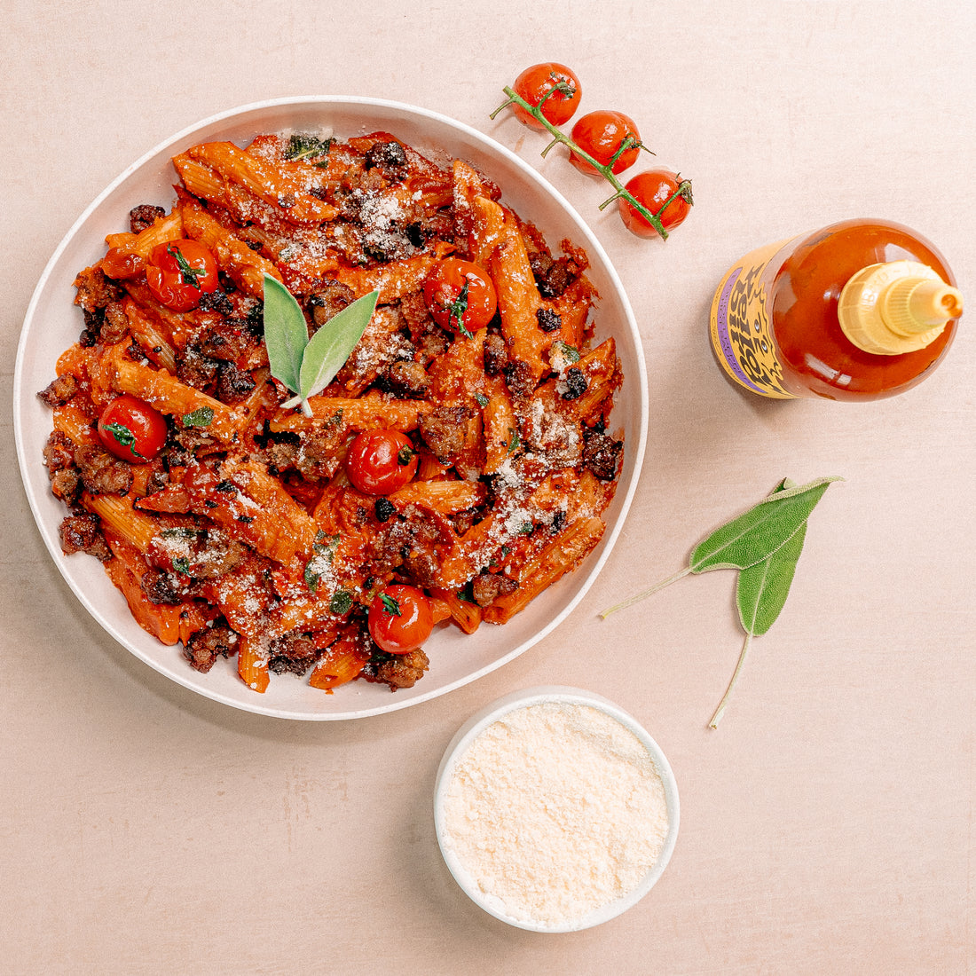 An overhead shot with a bowl of penne pasta in tomato sauce with sausage, blistered tomatoes, and sage. Next to a bowl of grated parmesan cheese and a bottle of Yellowbird Ghost Pepper Hot Sauce.