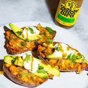 Three baked sweet potatoes with corn, avocado, sour cream cilantro, and a heavy drizzle of hot sauce, with a bottle of Yellowbird Serrano Hot Sauce in the background. 