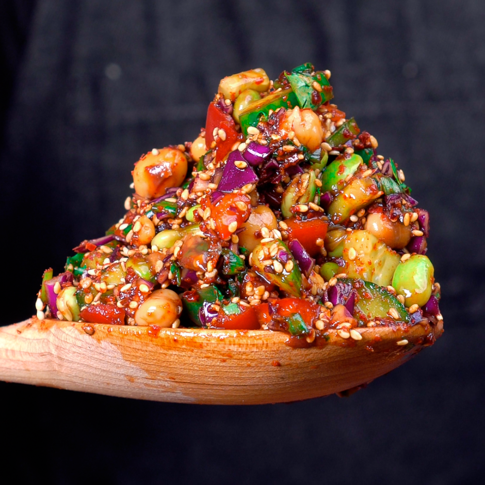 Close up of a heaping spoonful of Sriracha Sesame Edamame Salad, piled onto a wooden spoon. It looks fresh, saucy, and delicious with chunks of cucumber, bell pepper, chickpeas, edamame, cabbage, and coated in sauce and sesame seeds.