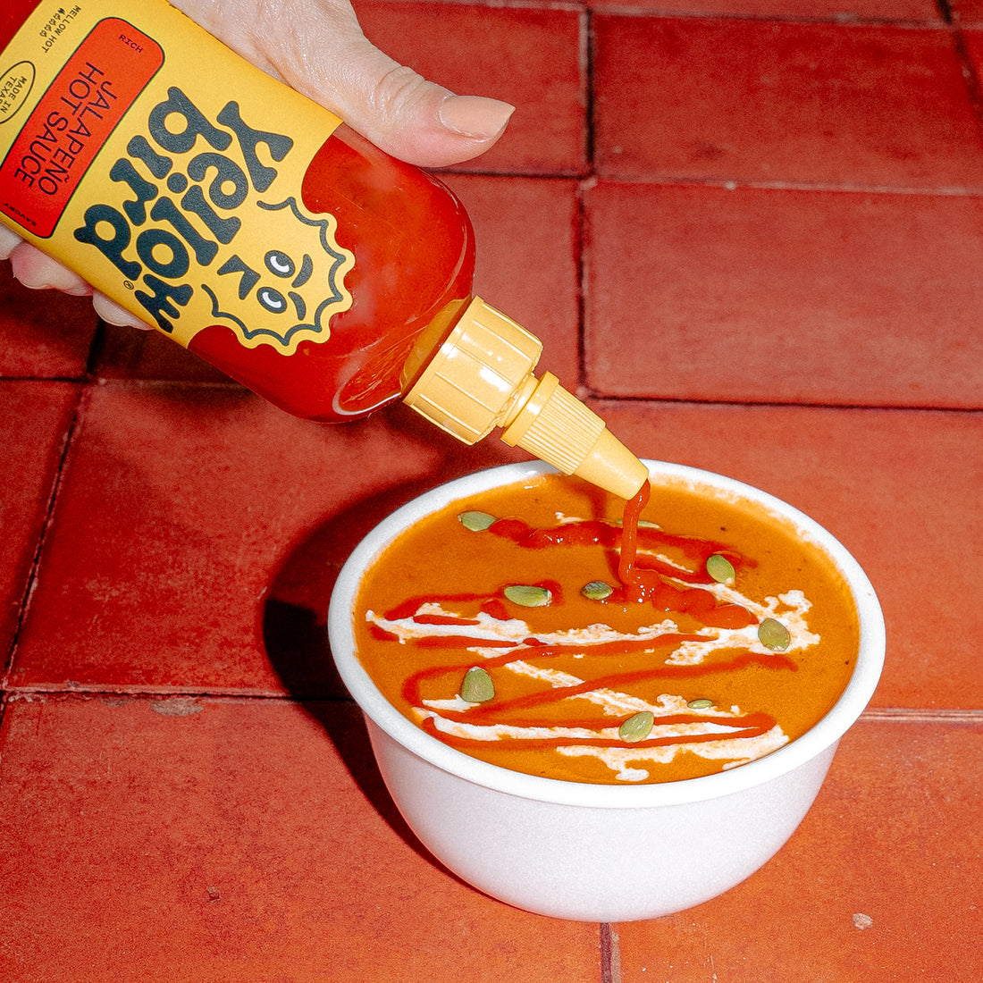 A bowl of tomato soup topped with heavy cream, toasted pumpkin seeds, and a generous drizzle of Yellowbird Jalapeño Hot Sauce.