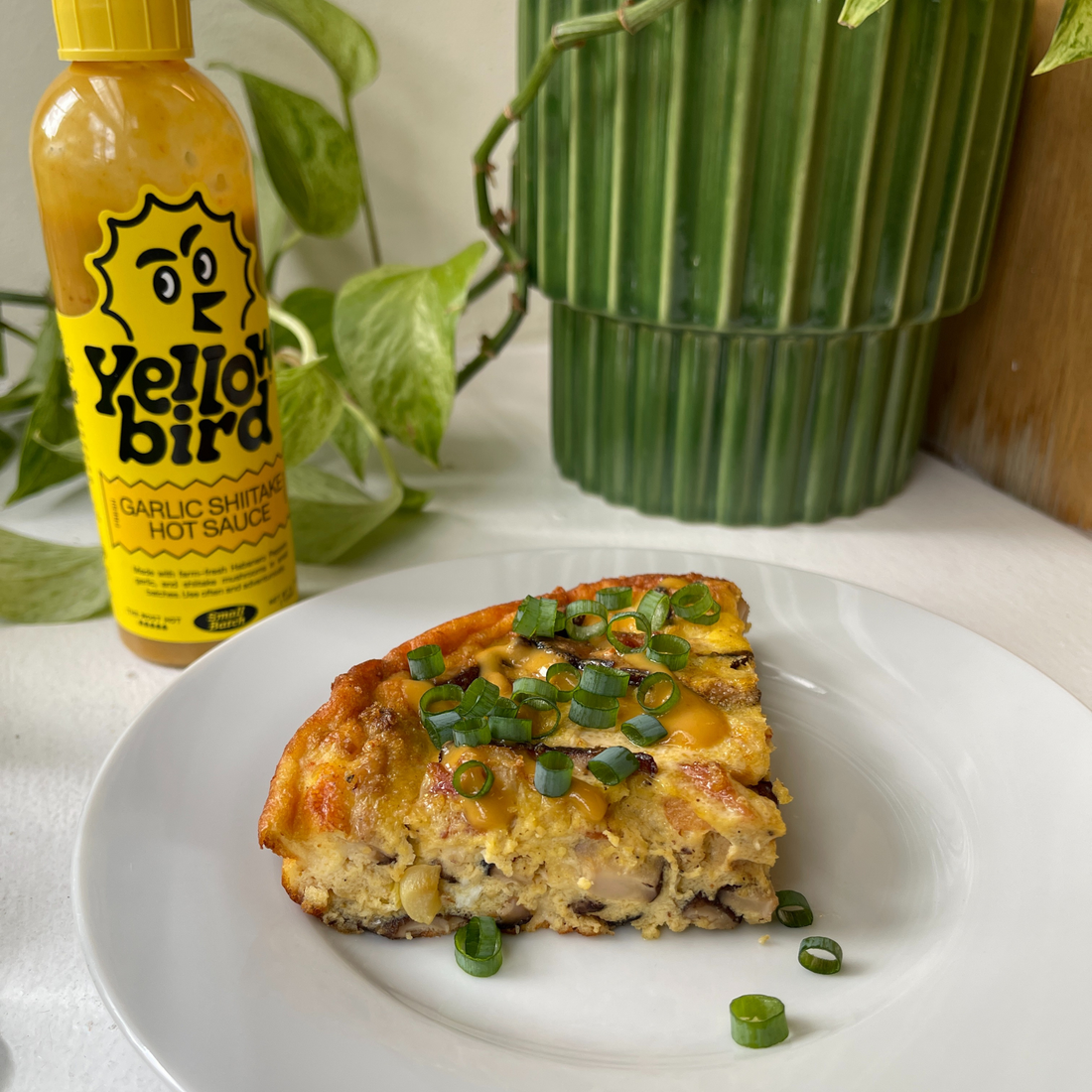 A slice of Frittata on a plate, garnished with scallions, next to a bottle of Yellowbird Garlic Shiitake Hot Sauce