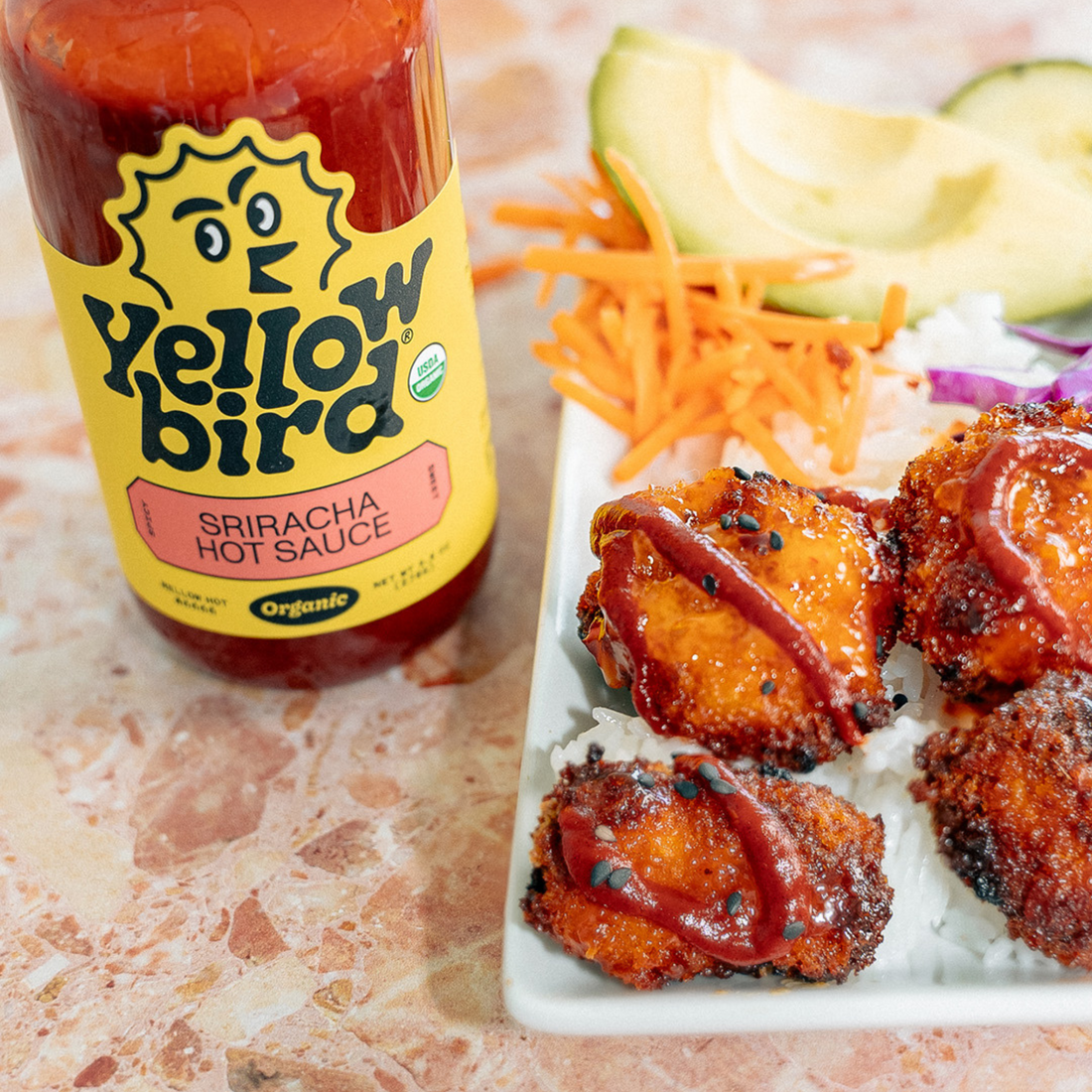 A bottle of Yellowbird Organic Sriracha next to fried chicken over rice coated in sauce, with carrots and avocado on the plate.