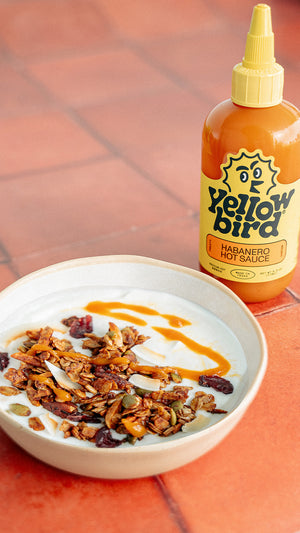 A bowl of yogurt with granola and Habanero Hot Sauce drizzled on it, with a bottle of Yellowbird Habanero Hot Sauce next to it.