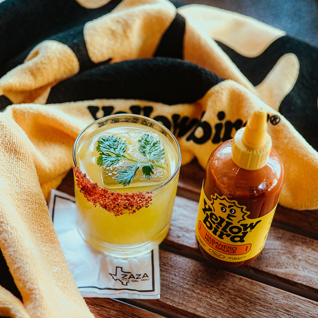 Spicy Sundown Cocktail in a glass, garnished with cilantro and rimmed with Tajin, next to a bottle of Yellowbird Habanero Hot Sauce